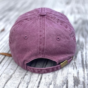 May include: A dusty rose baseball cap with a curved brim. The back of the cap features an adjustable strap with a brass-colored metal clasp. The cap is made of a soft, textured fabric.