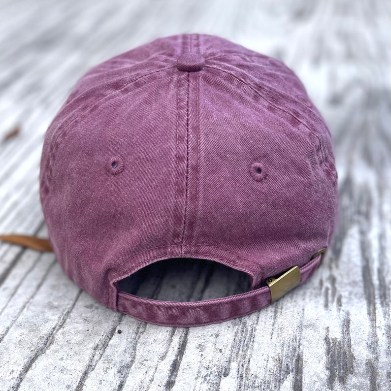 May include: A dusty rose baseball cap with a curved brim. The back of the cap features an adjustable strap with a brass-colored metal clasp. The cap is made of a soft, textured fabric.