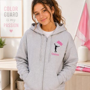May include: A light gray zip-up hoodie with a black graphic of a person holding a pink flag and the name "Chelsea" in pink. The framed artwork behind the person says "Color Guard is my Passion".