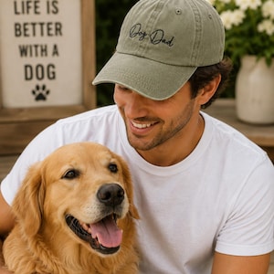 May include: An olive green baseball cap with the embroidered text "Dog Dad" in black script. A sign in the background reads "Life is better with a dog." A golden retriever dog is in the foreground.