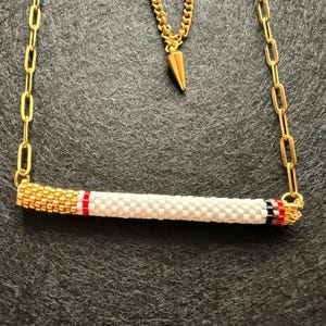 May include: A gold-toned chain necklace with a pendant and a beaded cigarette pendant. The cigarette pendant is white with gold, red, and black accents. The necklace is displayed on a dark gray surface.