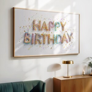 May include: A framed print with the words "HAPPY BIRTHDAY" spelled out in colorful, sprinkle-covered letters. The white background is speckled with confetti, and the frame is a light wood color. A festive birthday decoration.