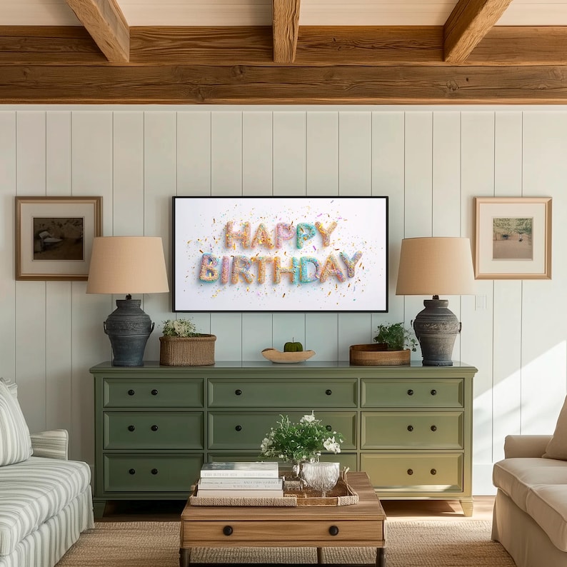 May include: A living room scene with a green dresser, two lamps, and a coffee table. A framed picture on the wall displays the words "HAPPY BIRTHDAY" in colorful letters. The room has a neutral color palette with wooden beams and light-colored walls.