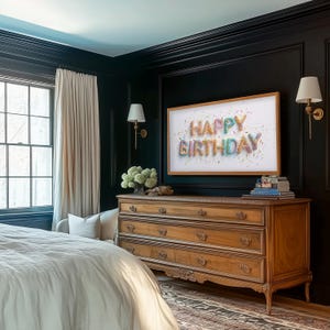 May include: A bedroom scene featuring a wooden dresser with four drawers, topped with books and a framed artwork that reads "HAPPY BIRTHDAY." The room has dark walls, a window with cream-colored curtains, and two sconces. The bed is covered with a white comforter.