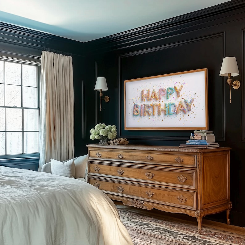 May include: A bedroom scene featuring a wooden dresser with four drawers, topped with books and a framed artwork that reads "HAPPY BIRTHDAY." The room has dark walls, a window with cream-colored curtains, and two sconces. The bed is covered with a white comforter.