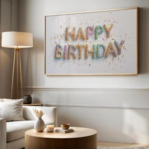 May include: A framed print with the words "HAPPY BIRTHDAY" in colorful, textured letters. The letters are in a 3D style, with confetti scattered around them. The frame is gold-colored, and the background is white.