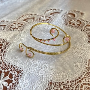 Può includere: Un bracciale dorato con perline rosa. Il bracciale presenta un design a spirale con curve delicate e piccole perline rosa. Il gioiello è posto su uno sfondo di pizzo bianco, evidenziando gli intricati dettagli del design.