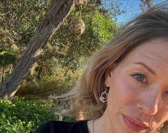 May include: A woman wearing silver hoop earrings with a small, textured design. The earrings are the focal point, set against a backdrop of green foliage, a tree trunk, and a clear blue sky. The woman is wearing a black shirt.