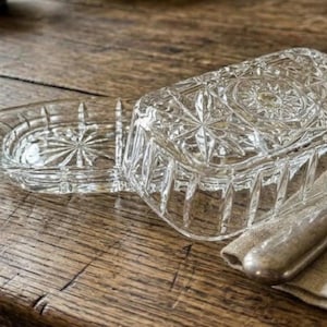 May include: A clear glass butter dish with a rectangular base and a matching lid, both featuring a pressed starburst pattern and vertical fluted sides. A metal butter knife with a worn handle rests on a folded beige linen napkin nearby.