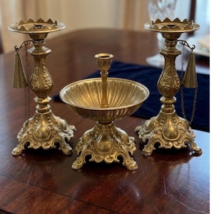 May include: A set of three antique brass candle holders. Two ornate candlesticks with bell-shaped pendants and a central bowl-shaped piece. The pieces have detailed floral and scrollwork designs, and are displayed on a dark wooden surface.