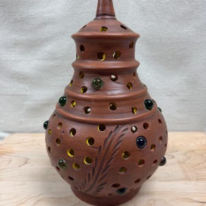 May include: A terracotta-colored ceramic lantern with a pointed lid, featuring numerous circular cutouts and embedded glass marbles in shades of green and blue. The surface is decorated with a carved leaf pattern and reveals a bright yellow interior.