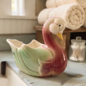 May include: A ceramic swan-shaped planter or dish featuring a glossy finish with a gradient of soft pink on the neck and chest transitioning into a pale green on the wings and body, with a yellow beak and a cream-colored interior basin.