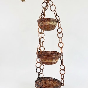 May include: A three-tiered hanging basket planter with woven brown and tan baskets suspended by a chain. The chain and the hook are a dark bronze color. The baskets are round and have a natural, rustic appearance.