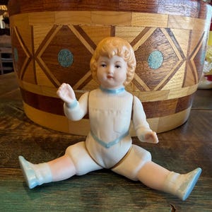 May include: A porcelain doll with blonde hair, wearing a white outfit with blue accents. The doll is posed with its legs spread. A wooden container with a geometric design is in the background.