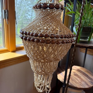 May include: A handcrafted chandelier with a tiered design, featuring cream-colored woven material and dark brown wooden beads. The chandelier is suspended from a ceiling fixture and is designed for indoor use. The light fixture is a decorative element.