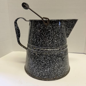 Może przedstawiać: Zabytkowy czarno-biały emaliowany dzbanek do kawy z dzióbkiem i uchwytem. Dzbanek ma druciany uchwyt z drewnianym uchwytem i zakrzywiony uchwyt z boku. Dzbanek ma około 20 cm wysokości.