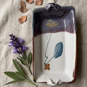 May include: Rectangular ceramic tray with a purple and white glaze. The tray features a painted blue and brown floral design. The edges are a dark brown color. The tray has a handle on each end.