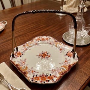 May include: A decorative serving tray with a woven handle. The tray features a floral design in shades of orange, red, and gold against a cream background. The handle is brown and the tray is placed on a wooden table.