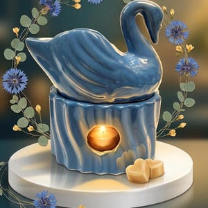 May include: A blue ceramic swan-shaped oil warmer with a candle inside. The warmer sits on a white circular base, with two heart-shaped wax melts. The swan is surrounded by a floral wreath of blue flowers and green leaves.