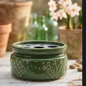 May include: A dark green ceramic flower frog with a round shape and a series of holes on top. The sides are decorated with embossed floral designs. The background includes other ceramic pots, green glass bottles, and potted flowers.