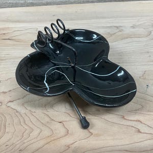 May include: A black, three-sectioned dish with white accent lines and a metal stand. The dish has a glossy finish and a decorative metal coil on top. The stand has a small, black, rounded foot.