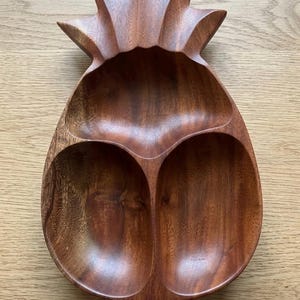 Könnte beinhalten: Ein hölzernes Ananas-Serviertablett mit drei Fächern. Das Tablett ist aus dunkelbraunem Holz gefertigt und hat eine natürliche Holzmaserung. Die Oberseite des Tabletts ist wie eine Ananaskrone geformt.