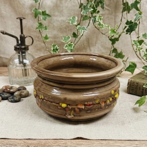 May include: A brown ceramic planter with a decorative band of yellow, orange, and red accents. The planter has a wide, open top and a slightly textured surface. A vintage glass mister and decorative stones are in the background.