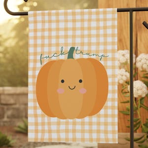 Può includere: Una bandiera da giardino con un design a forma di zucca dei cartoni animati e la scritta "fuck trump". La zucca arancione con un volto sorridente e un gambo verde è posta su un motivo a quadri vichy giallo e bianco.