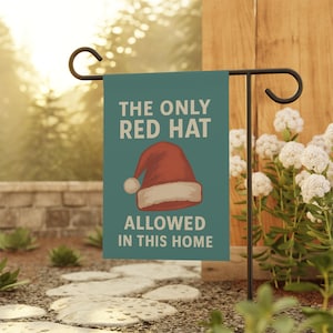 May include: A teal garden flag with the text "THE ONLY RED HAT ALLOWED IN THIS HOME" and a graphic of a red Santa hat. The flag is displayed on a black metal stand in a garden setting.