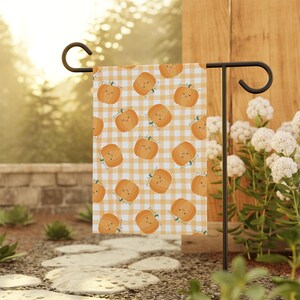 Puede incluir: Una bandera de jardín con un patrón repetido de calabazas naranjas sonrientes sobre un fondo de cuadros amarillos y blancos. La bandera se muestra en un soporte de metal negro en un jardín, con vegetación y flores blancas en el fondo.