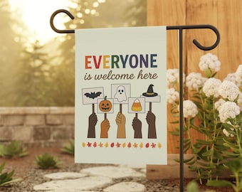 May include: A beige garden flag with the text "EVERYONE is welcome here" in orange, blue, and green. The flag features illustrations of a bat, pumpkin, ghost, witch hat, and candy corn. The flag is held by a black metal stand.