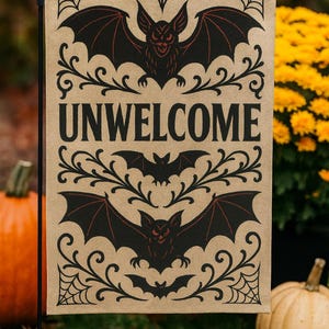 Könnte beinhalten: Eine Halloween-Gartenflagge mit beigem Hintergrund zeigt schwarze Fledermäuse und das Wort "UNWELCOME". Die Fledermäuse haben rote Akzente und sind von dekorativen Schnörkeln und Spinnweben umgeben. Eine festliche Außendekoration.