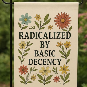 Op de afbeelding: Een beige tuinvaandel met de tekst "RADICALIZED BY BASIC DECENCY" in het zwart. De vlag is versierd met kleurrijke bloemen in tinten blauw, oranje en geel, met groene bladeren. De vlag hangt aan een zwarte metalen standaard.