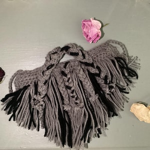 May include: Handmade gray and black crochet beard with braided details and fringe. The beard is adorned with black accents and fringe. Dried roses in pink, red, and white are scattered around the beard.