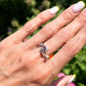 Puede incluir: Un anillo de plata con un diseño de peces koi. El anillo tiene dos peces, cada uno con aletas y escamas detalladas, nadando en direcciones opuestas. Los peces tienen detalles en azul oscuro. El anillo se lleva en una mano.