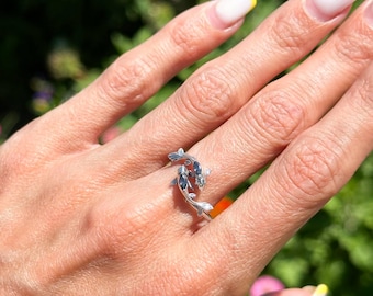 May include: A silver ring featuring a koi fish design. The ring has two fish, each with detailed fins and scales, swimming in opposite directions. The fish have dark blue accents. The ring is worn on a hand with a natural background.