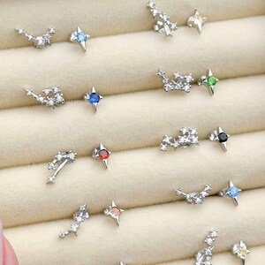 May include: A collection of silver-toned stud earrings with various designs. Some earrings feature clusters of small, clear stones, while others have a star shape with a colored gemstone center. The gemstones are in shades of blue, red, green, and black.