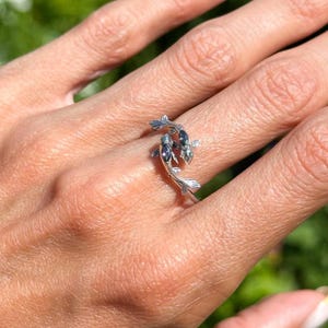 Puede incluir: Un anillo de plata con un diseño de dos carpas koi. Los peces tienen detalles de aletas y acentos oscuros, formando el anillo. El anillo se muestra en una mano, con un fondo verde borroso. El anillo está hecho de plata.
