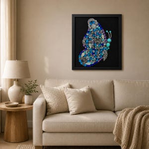 Könnte beinhalten: Ein quadratisches Wandbild mit einem Schmetterlingsmosaik aus verschiedenen blauen, türkisfarbenen und silberfarbenen Schmuckstücken, Perlen und Strasssteinen, angeordnet auf einem schwarzen Hintergrund in einem schlichten schwarzen Rahmen.
