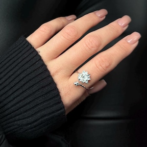 May include: A silver-colored ring featuring a flower design with a central stone. The ring wraps around the finger with an open design. The ring is worn on a hand with manicured nails, against a black background.
