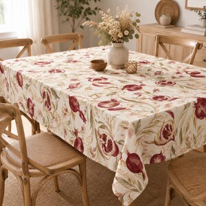 May include: A rectangular tablecloth featuring a repeating pattern of deep red pomegranates and soft green foliage on a cream-colored background. The fabric drapes over a dining table surrounded by wooden chairs with woven seats.