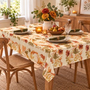 May include: A rectangular tablecloth with a cream background featuring a repeating pattern of orange, red, and green autumn leaves and swirling scrollwork. The table is set with white plates, green napkins, crystal glasses, and a bowl of fruit.