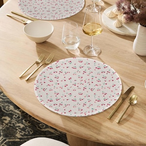 May include: White placemats with a repeating pattern of pink cherries and green leaves. The placemats are round and sit on a wooden table, alongside gold-colored cutlery, a white bowl, and glassware.