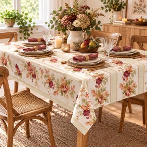 May include: A rectangular dining table covered with a cream-colored tablecloth featuring a floral pattern of red, orange, and green leaves. The table is set with white plates, woven placemats, pink cloth napkins, and crystal glasses.