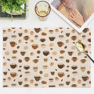 May include: Beige placemat with a repeating pattern of coffee cups, beans, and pastries in shades of brown. A gold spoon rests on the right side. A glass and a plant are visible in the background.