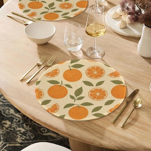 May include: Round placemats with an orange fruit pattern. The placemats feature whole and sliced oranges with green leaves on a cream background. Gold-colored cutlery and a white bowl are on the wooden table.