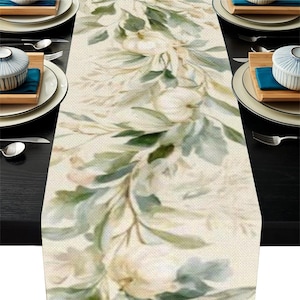 May include: A rectangular table runner featuring a printed pattern of soft green and sage leaves, vines, and white pumpkins on a textured beige fabric background. The runner is displayed on a dark surface with white plates and silverware.