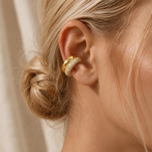 Könnte beinhalten: Ein goldener Ohrklemme mit Doppelband-Design. Das obere Band ist mit kleinen, funkelnden Kristallen besetzt, während das untere Band glatt ist. Der Schmuck wird am Ohr getragen und zeigt seinen eleganten und modernen Stil.