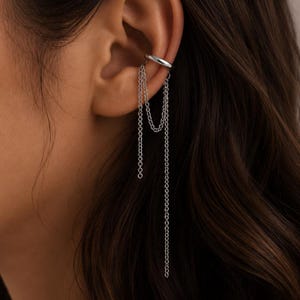May include: Silver ear cuff featuring a dangling chain design. The cuff wraps around the ear cartilage, with multiple delicate chains cascading downwards. The jewellery is made of a shiny, metallic material.