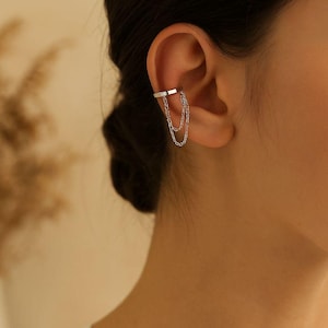May include: Silver ear cuff with two delicate chain dangles. The cuff wraps around the ear's cartilage, creating a modern and minimalist look. The chains add movement and a touch of elegance to the ear jewelry.